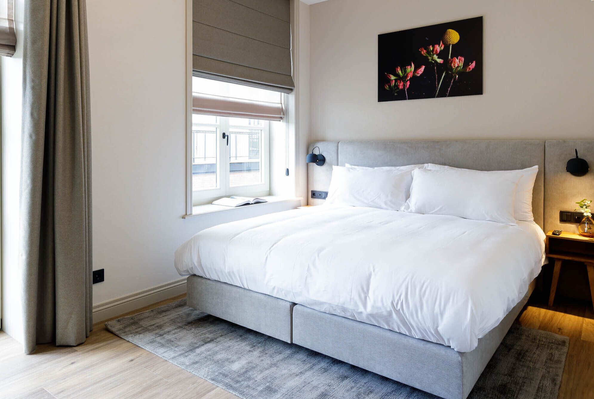Ein modernes Schlafzimmer mit einem grauen Bett, weißer Bettwäsche, einem grauen Kopfteil, einem Nachttisch mit Lampe und einem Fenster mit Vorhängen.