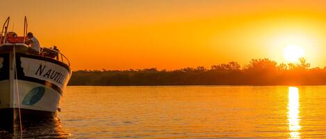 A boat named "NAUTILLA" with a person on deck, calm water, and a sunset in the background.