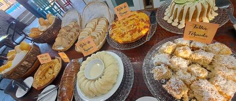 Mesa de buffet com diversos pratos identificados, incluindo pão, bolos e sobremesas.