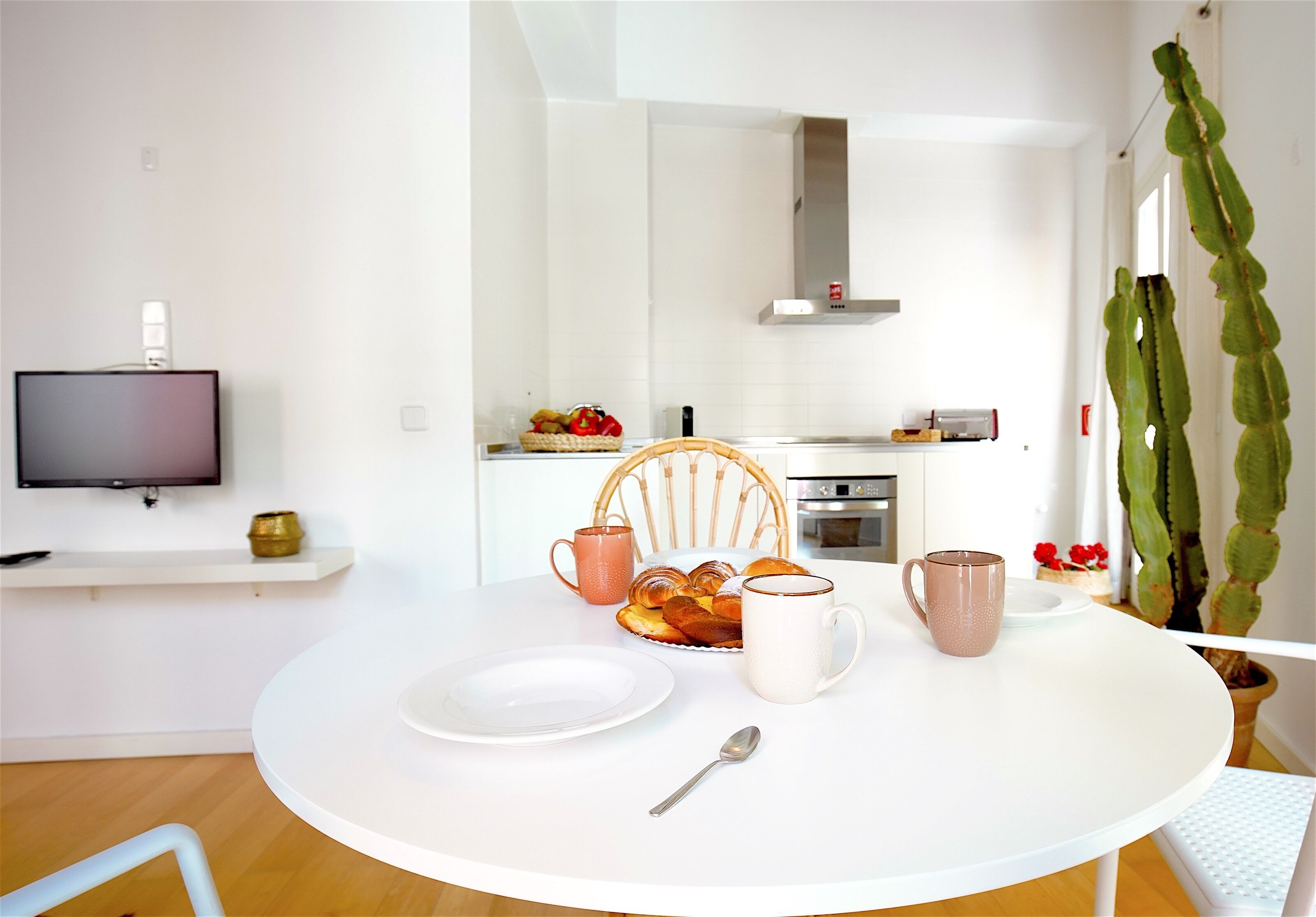 A dining area with a round table, white plates, a mug, and a bowl of pastries. A television is mounted on the wall, and there is a chair with a cushion.