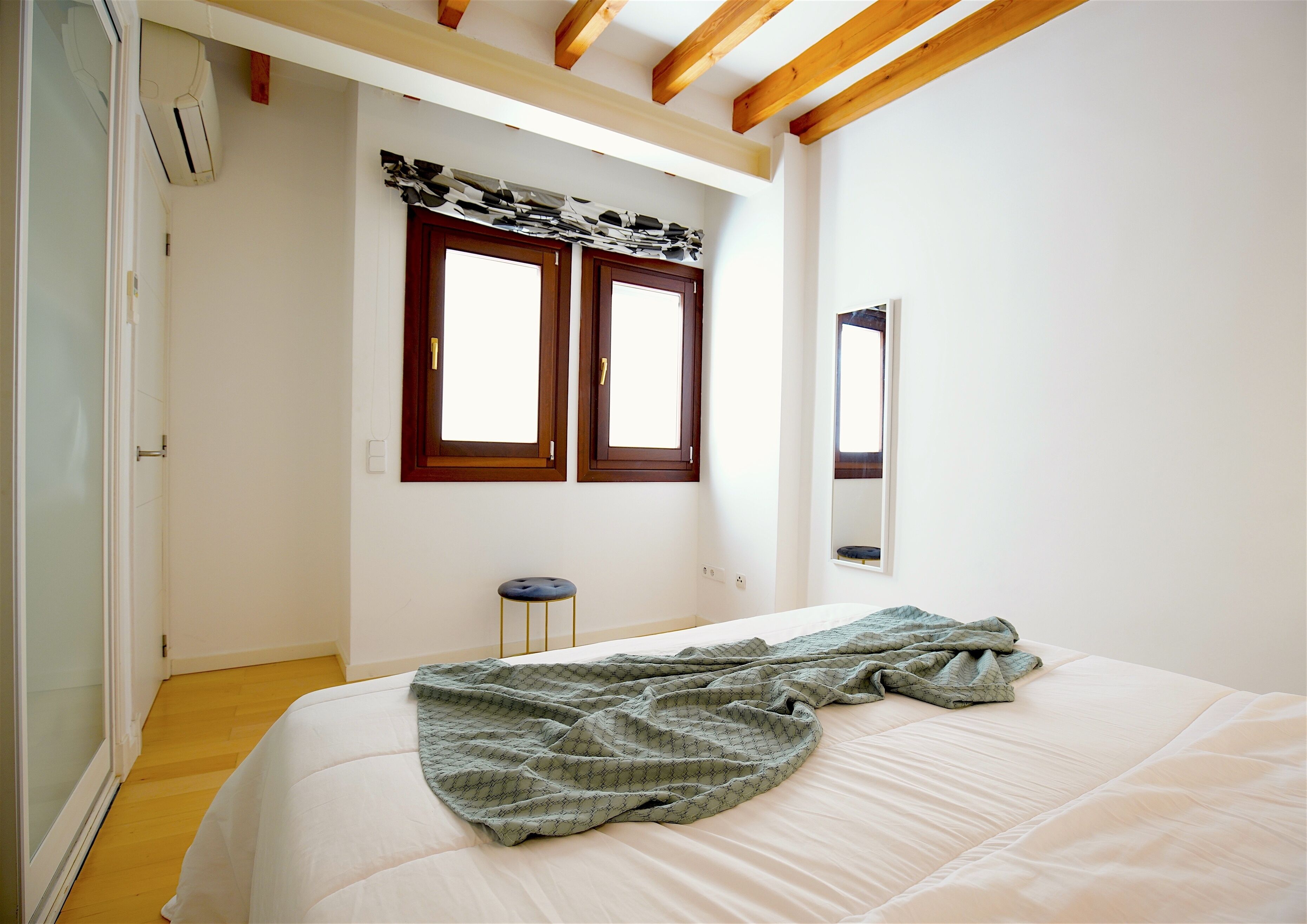 Een slaapkamer met een bed, een raam met gordijnen, een kruk en een spiegel aan de muur.