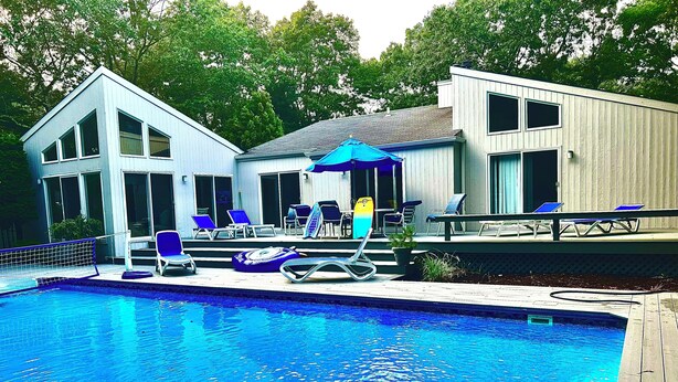 A swimming pool with lounge chairs and umbrellas, surrounded by a wooden deck and a building with large windows. - East Hampton SPECIAL (East Hampton)