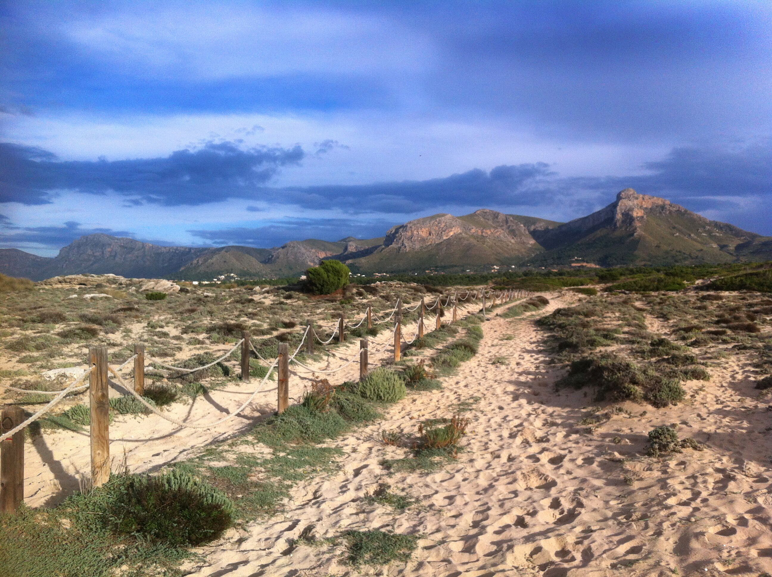A sandy path with wooden posts and ropes, leading towards a mountainous landscape.