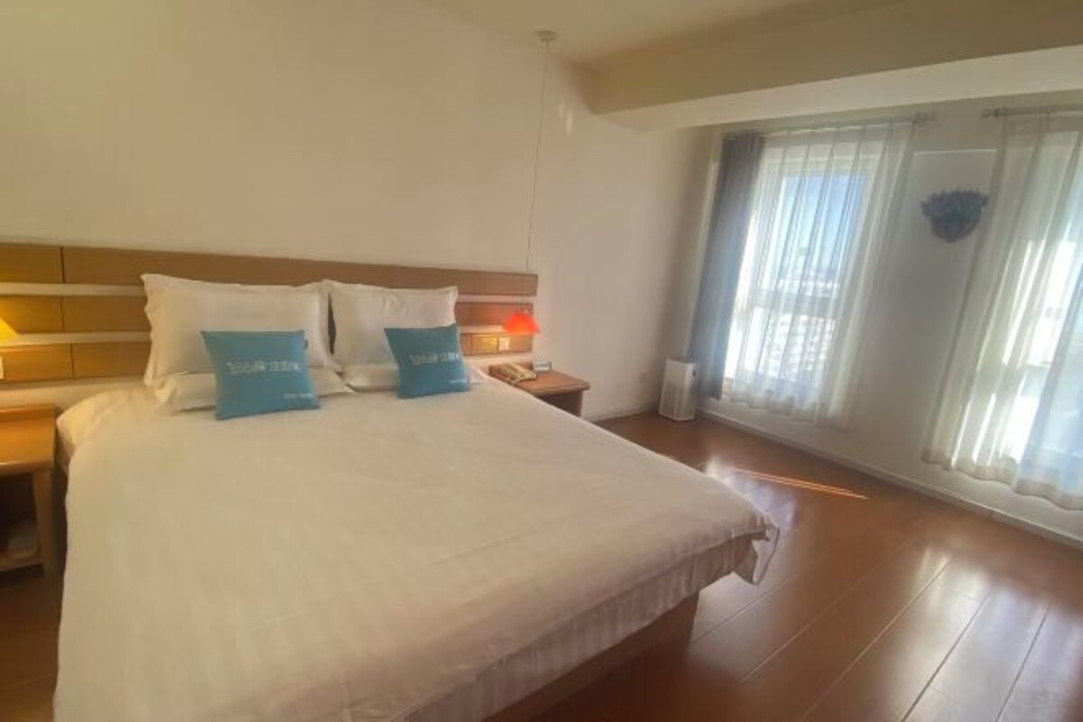 Camera d'albergo con un letto grande, un comodino, una lampada e vista sull'esterno attraverso una finestra con tende leggere.