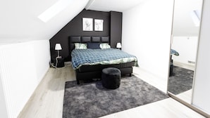Ein modernes Schlafzimmer mit schrägen Wänden, einem schwarzen Bett mit blau-grün kariertem Überwurf, zwei Wandleuchten und einem schwarzen Ottomanen.