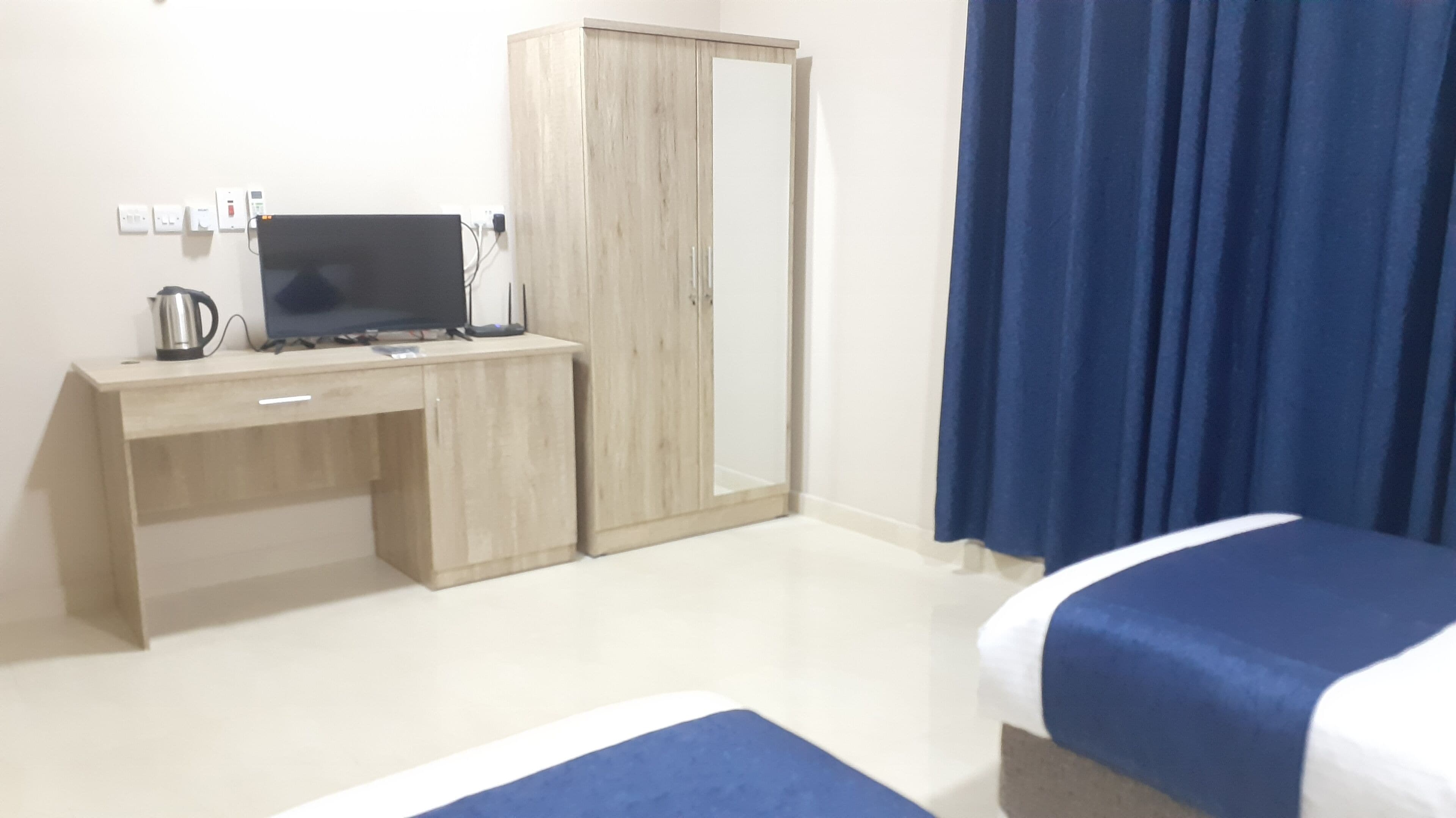 A hotel room with a wooden desk, a computer, a kettle, a wardrobe, and a bed with blue bedding.