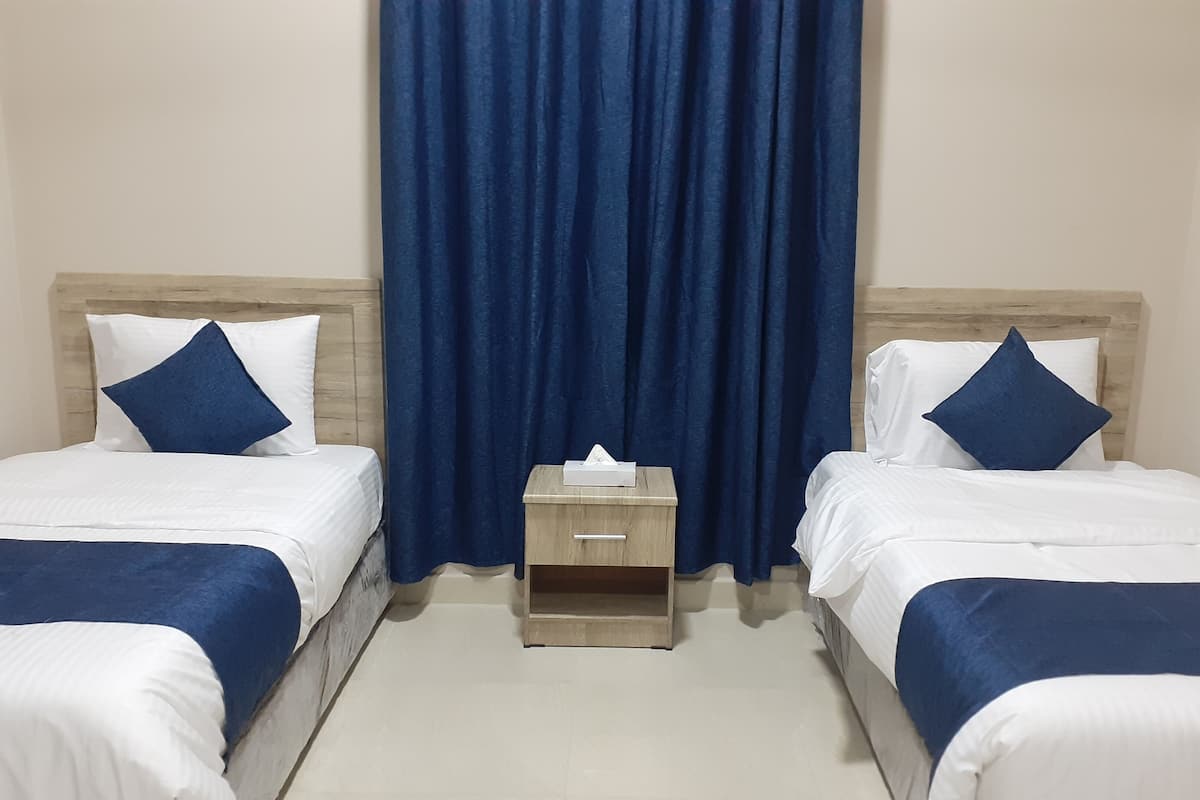 A hotel room with two single beds, each with a wooden headboard, white bedding, and blue pillows. A small wooden nightstand with a tissue box is between the beds.