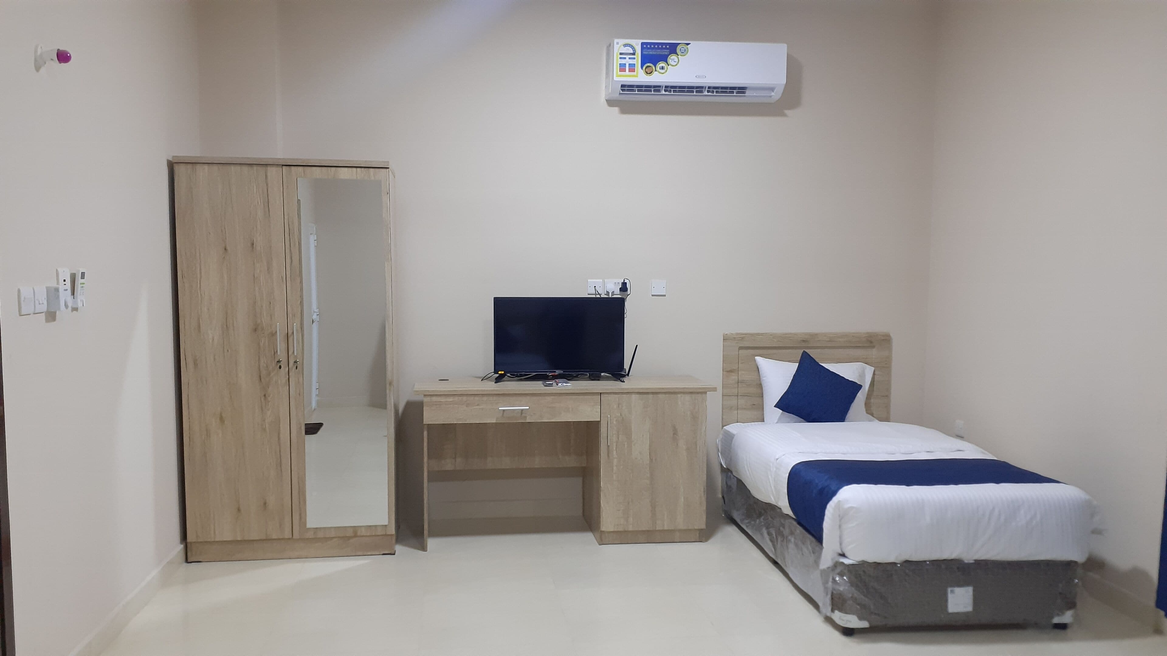 A bedroom with a wooden wardrobe, a desk with a computer, and a bed with a blue pillow.