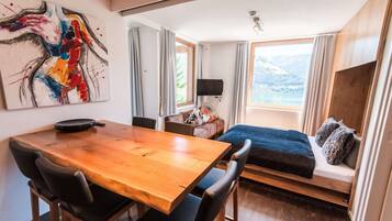 Een moderne kamer met een houten eettafel, stoelen, een bed met uitzicht, een flatscreen televisie en een groot abstract schilderij aan de muur.