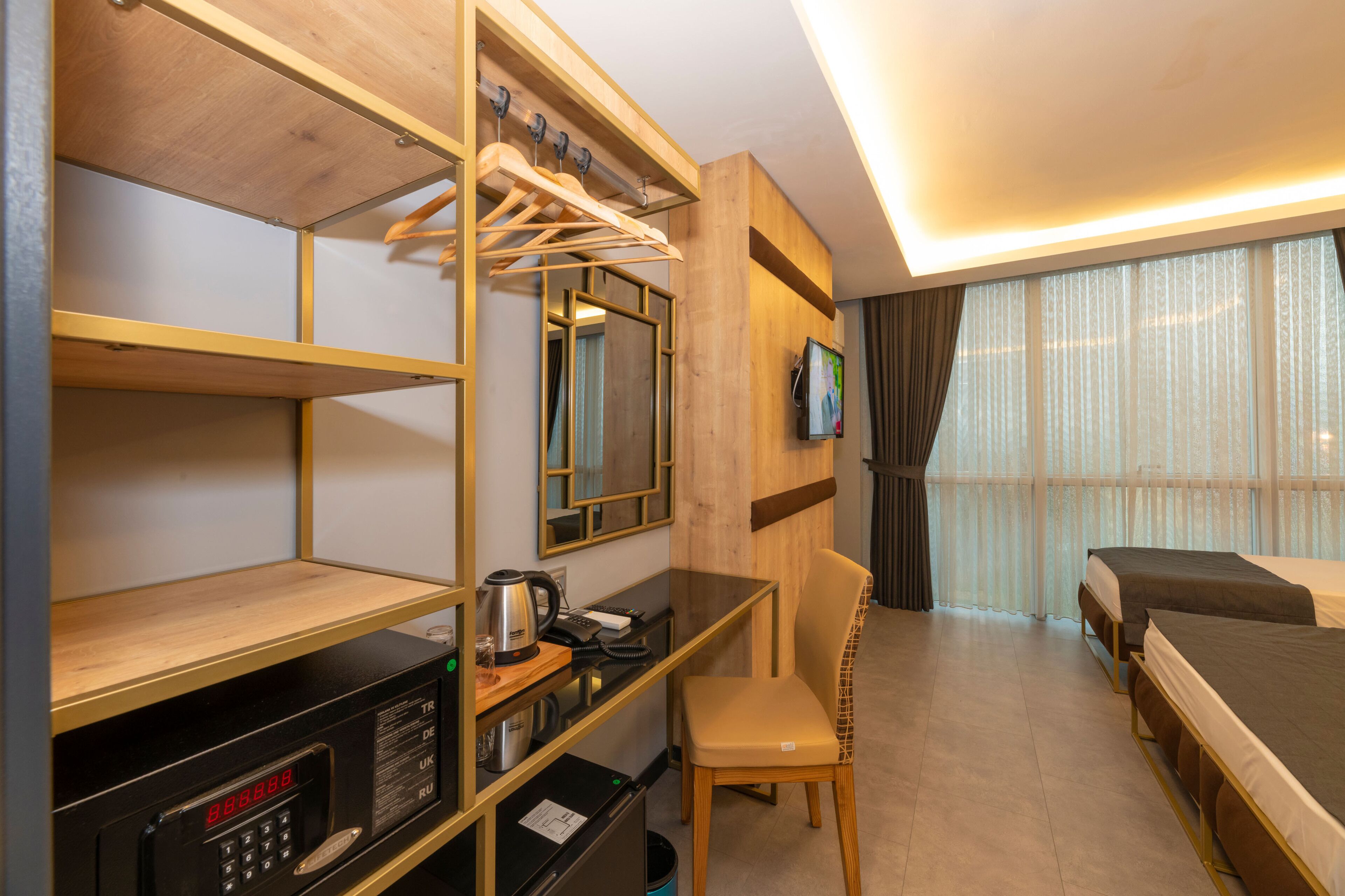 A hotel room with a bed, a desk with a kettle, a chair, a TV, and a wardrobe with hanging clothes.