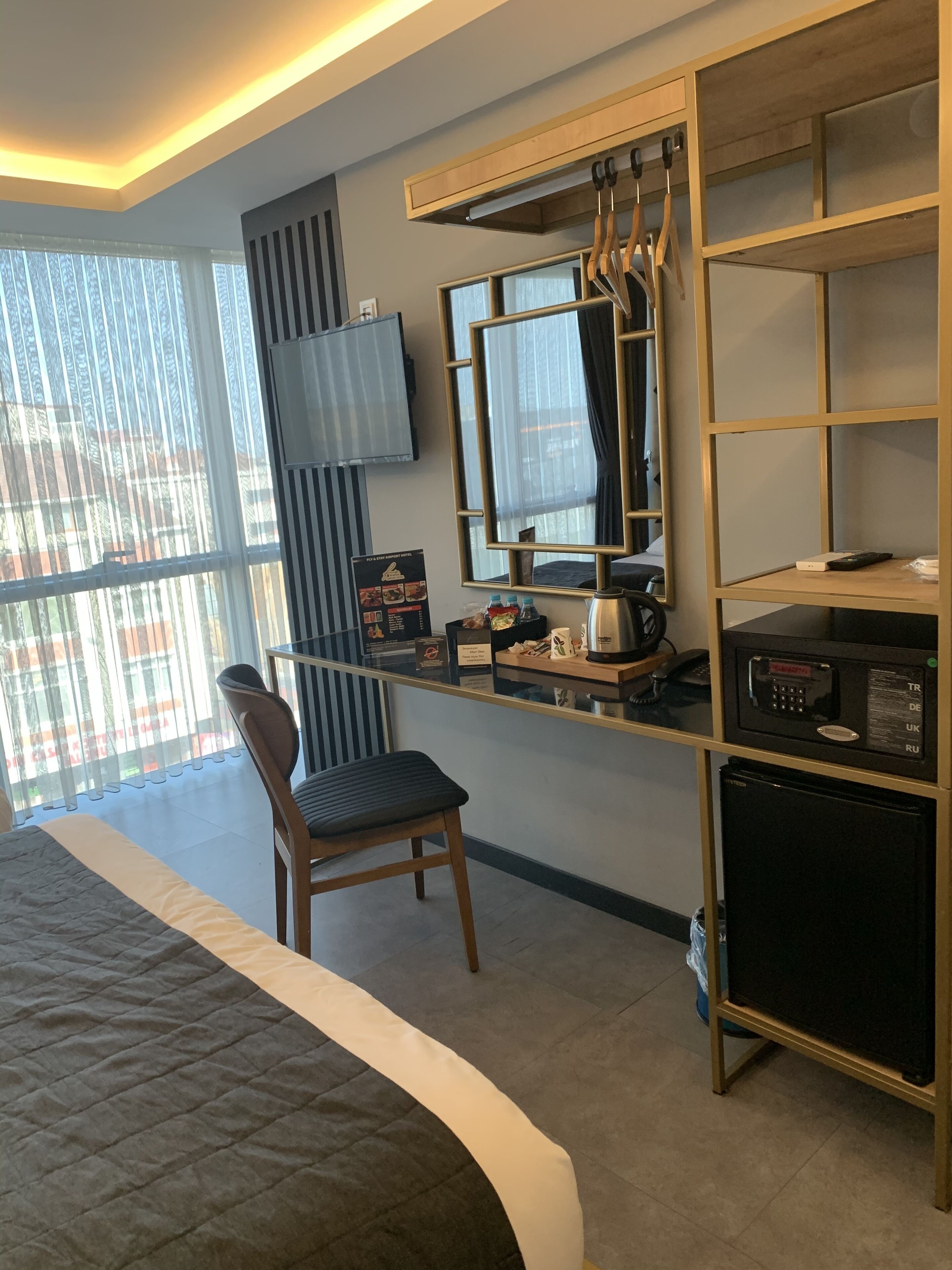 A modern hotel room with a bed, a desk with a computer, a chair, a TV, and a minibar.