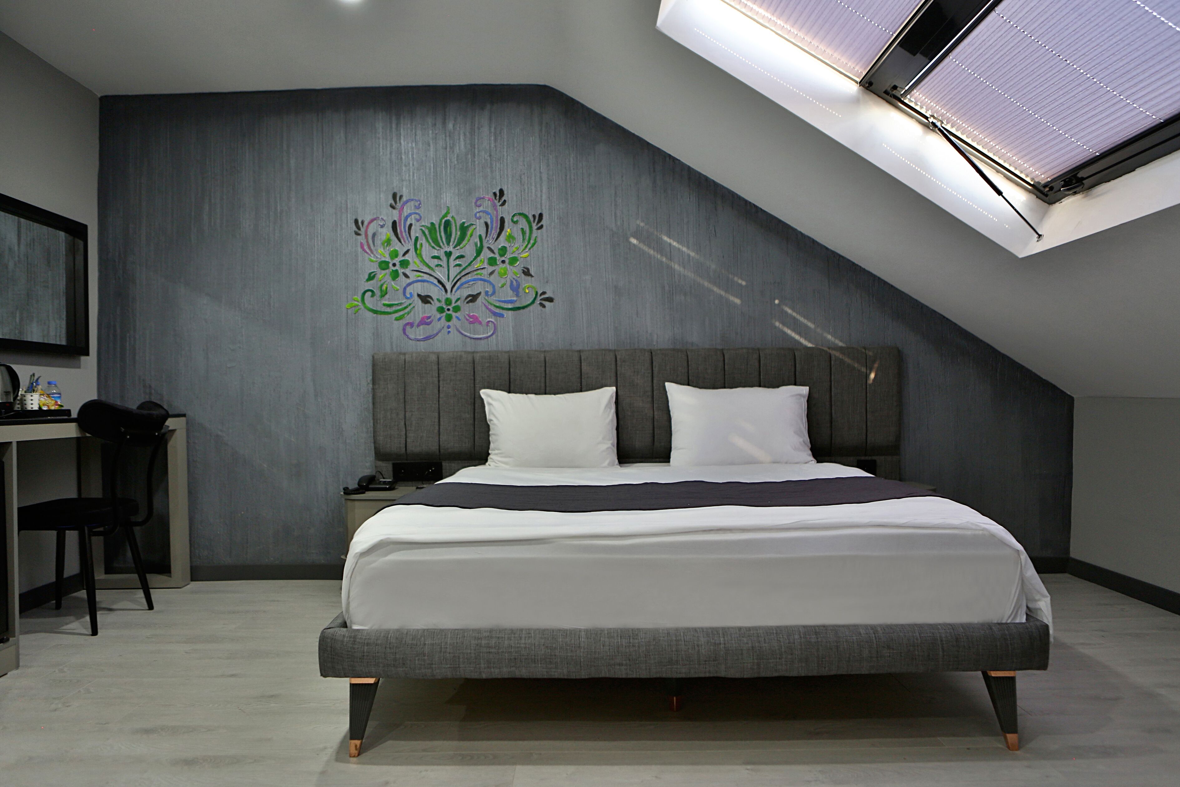 A modern bedroom with a skylight, a bed with white linens, a grey headboard, and a desk with a chair.