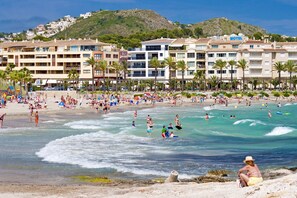 A beachfront hotel with multiple floors, a beach in the foreground, and a mountainous backdrop. - 3380 Villa Portichol Mar Flores (Moraira)