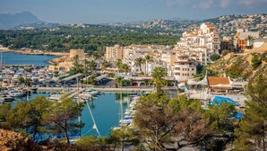 A coastal town with a marina, buildings with Mediterranean architecture, and a mountainous backdrop.