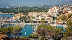 A coastal town with a marina, buildings with Mediterranean architecture, and a mountainous backdrop.