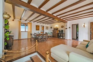 Een ruime woonkamer met een hoge plafond, houten balken, een eettafel met stoelen, een bank en een televisie.