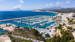 A marina with numerous boats docked, surrounded by a coastal town with buildings and a mountainous backdrop.