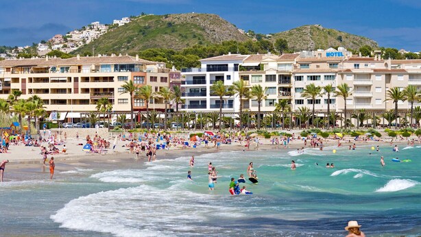 A beachfront hotel with multiple floors, a beach in the foreground, and a mountainous backdrop.