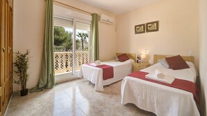 A hotel room with two beds, a sliding glass door to a balcony, a potted plant, and a wall-mounted air conditioner.