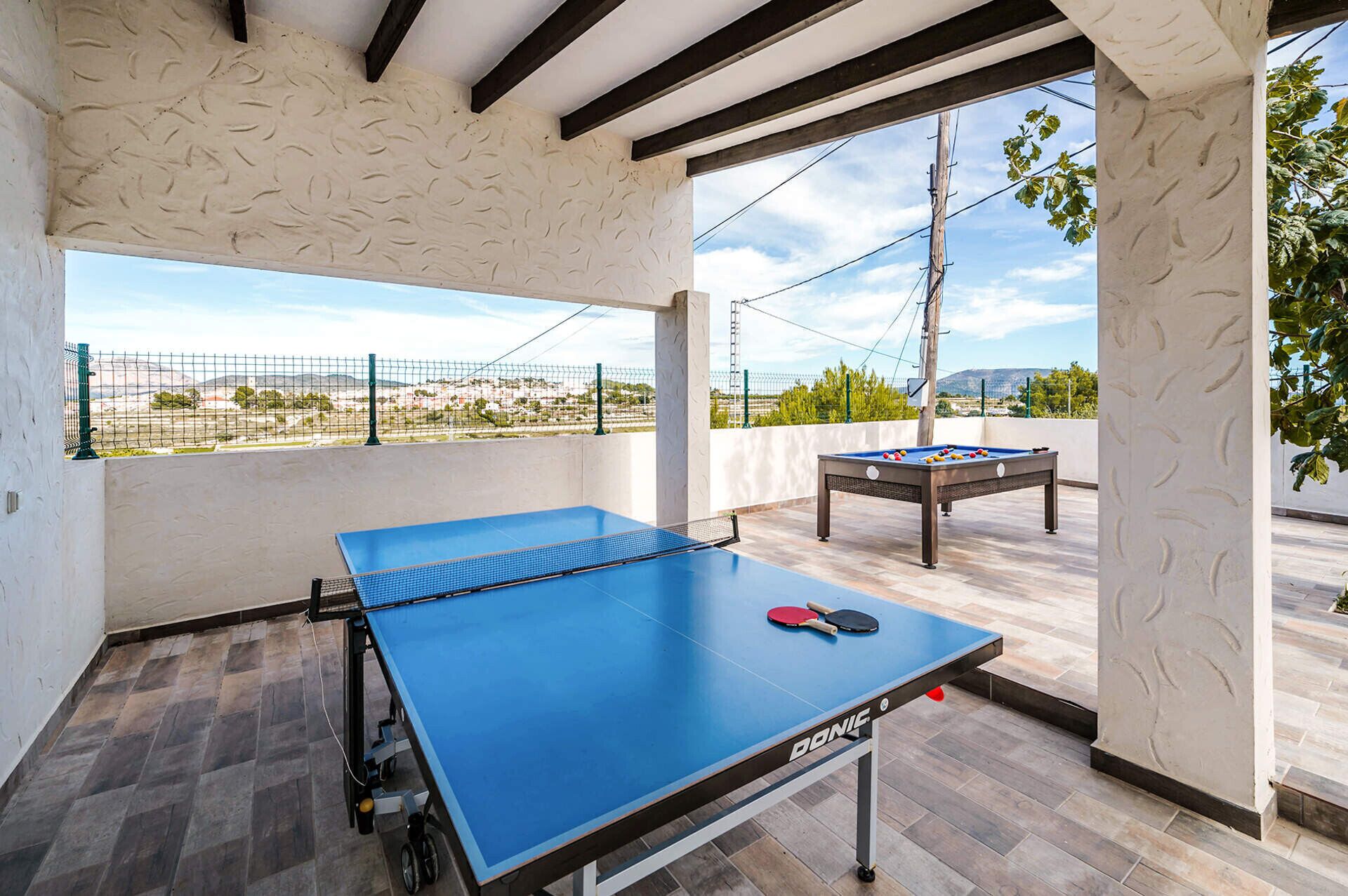 A ping pong table with a net, a pool table, and a table tennis paddle set up on an outdoor patio with a view of a distant landscape.
