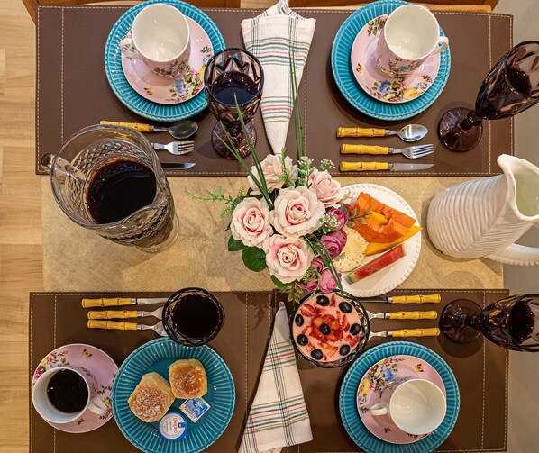 A table set for a meal with floral tablecloths, a centerpiece of roses, and various dishes including bread, fruit, and a pitcher. - Pousada Flor de Lótus (Ubatuba)