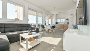 A modern living room with a grey sofa, a white coffee table, and a kitchen area with cabinets and a dining table. - Funtastic C Apartment with Pool and Terrace (Novalja)