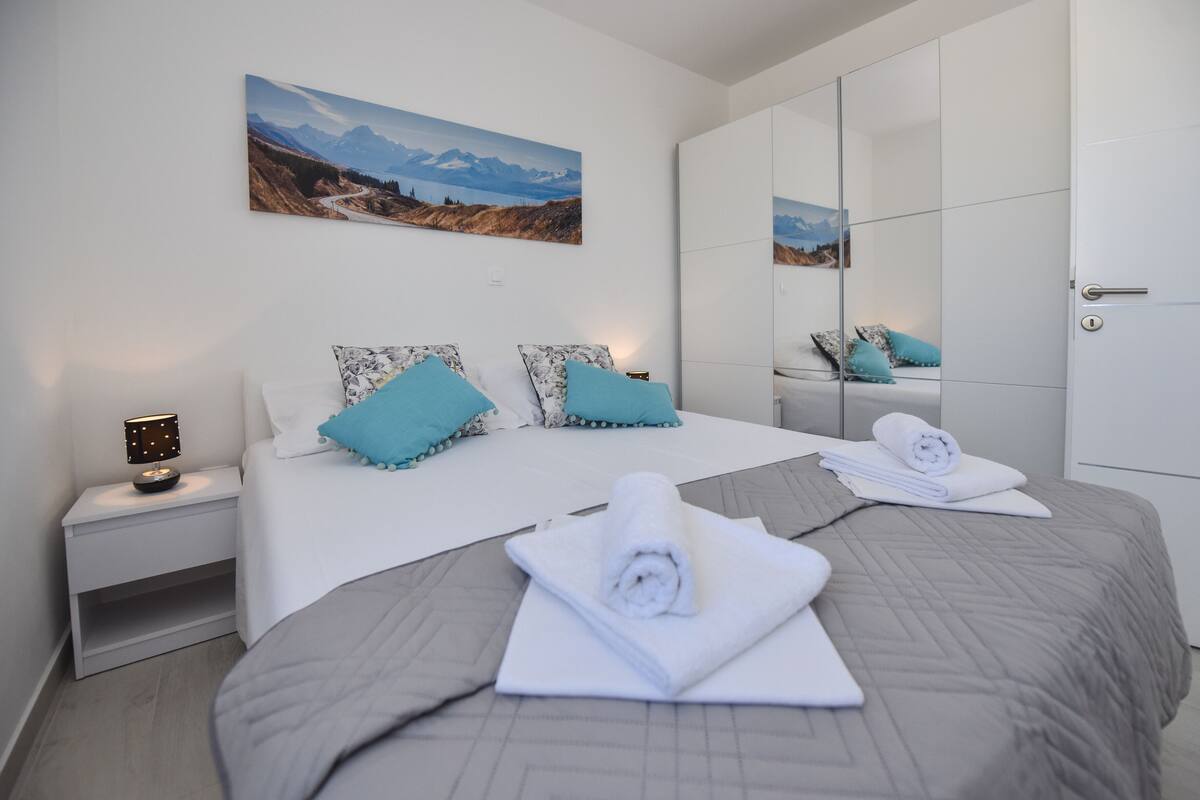 A modern hotel room with a large bed, a nightstand with a lamp, a painting on the wall, and a wardrobe with a mirror.