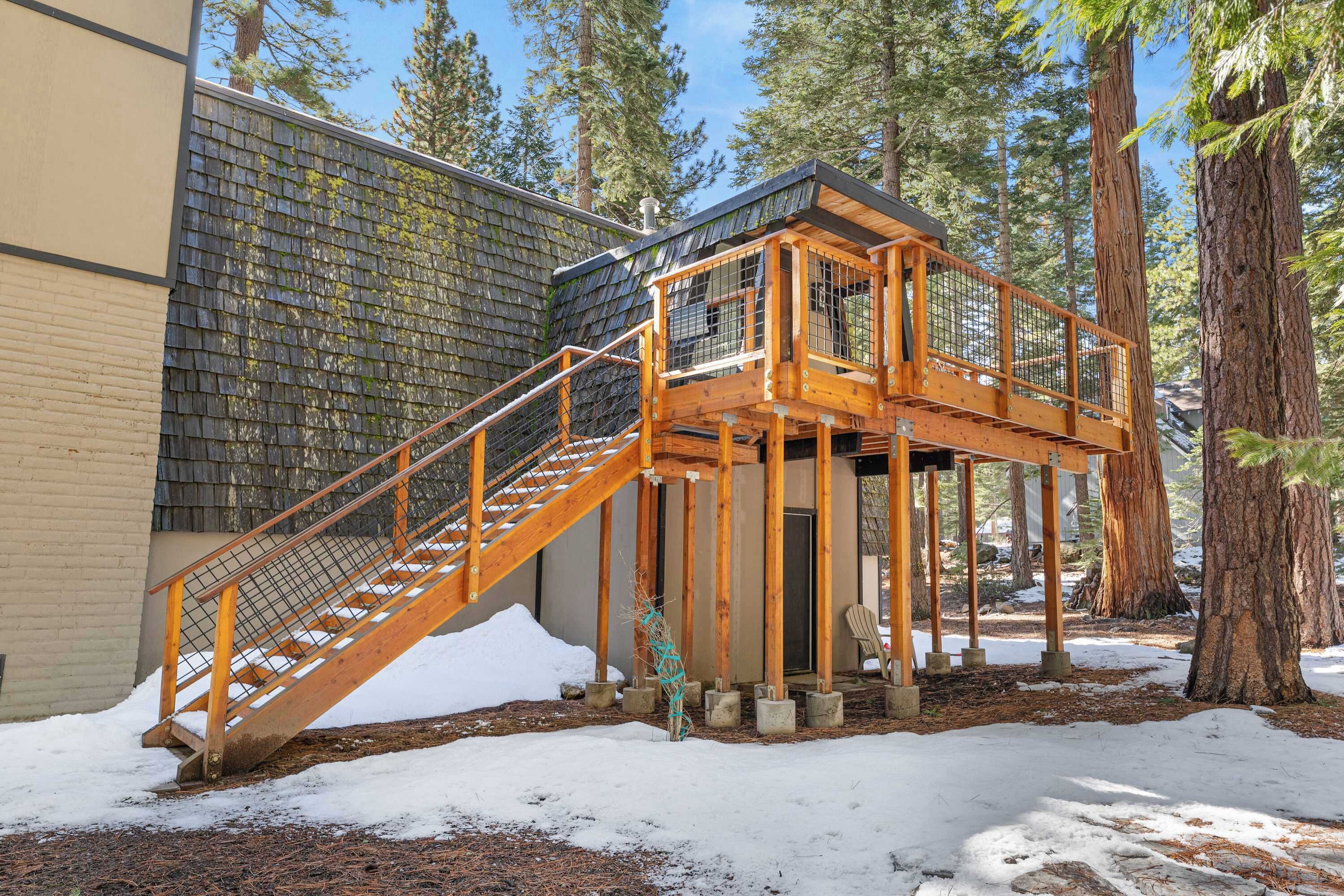 A wooden staircase leading to a deck with a metal railing, surrounded by trees and snow.