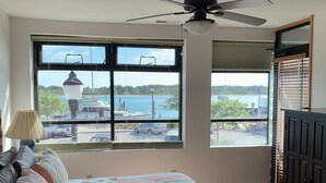 Quarto com cama, ventoinha no teto, janela com vista para um corpo de água e porta.