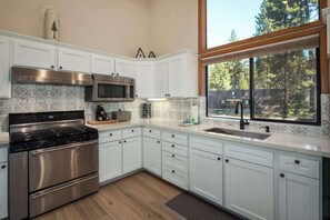A modern kitchen with white cabinets, stainless steel appliances, and a large window offering a view of trees.