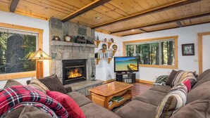 A cozy living room with a stone fireplace, a television, and a large window overlooking a forest. - Hot Tub with Forested Views plus Garage in Quiet Neighborhood (Tahoma)