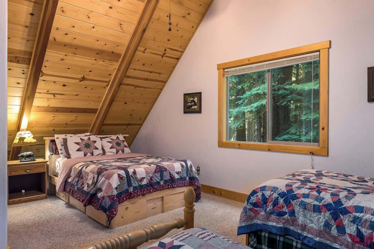 Une chambre avec un plafond en bois, une grande fenêtre donnant sur la forêt, un lit recouvert d’une courtepointe et une table de chevet avec une lampe.