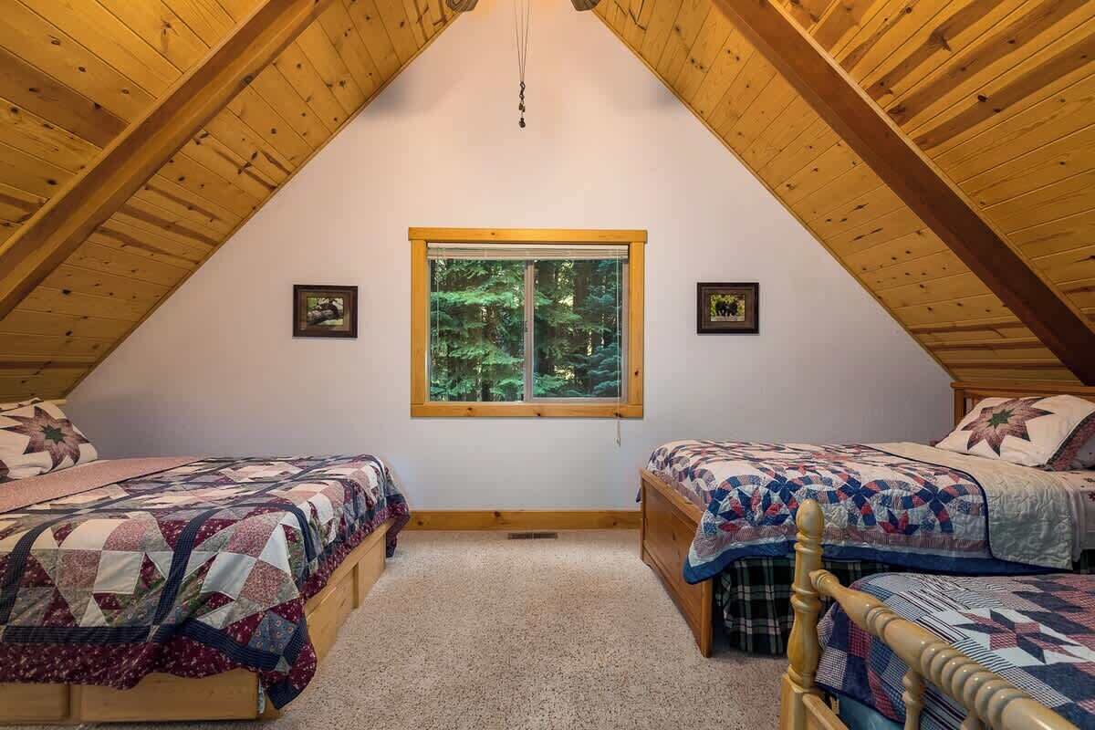 A bedroom with two beds, wooden beams, a window with a forest view, and a carpeted floor.