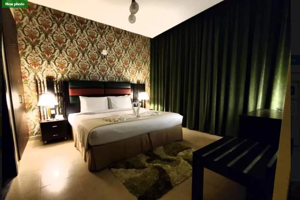 Quarto de hotel com uma cama grande, duas mesas de cabeceira com luminárias, cortina verde e papel de parede estampado.