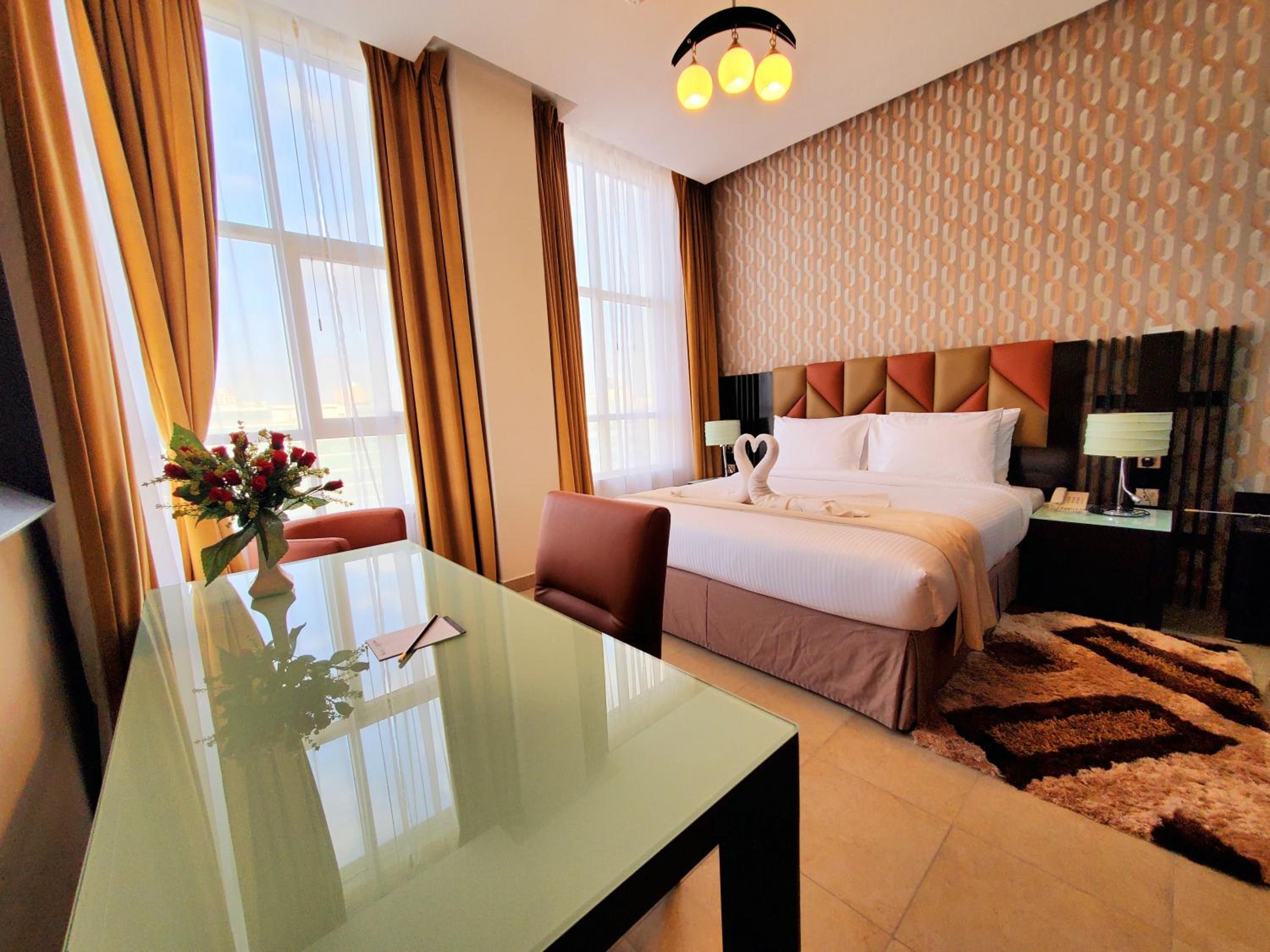 A hotel room with a bed, a glass table, a desk, a chair, a vase with flowers, and a patterned wall.