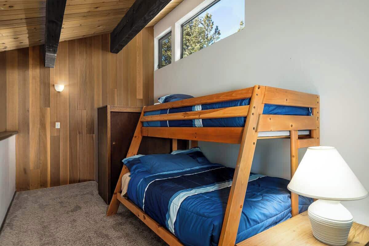 A bunk bed room with a wooden ceiling, a window, and a lamp on the nightstand.