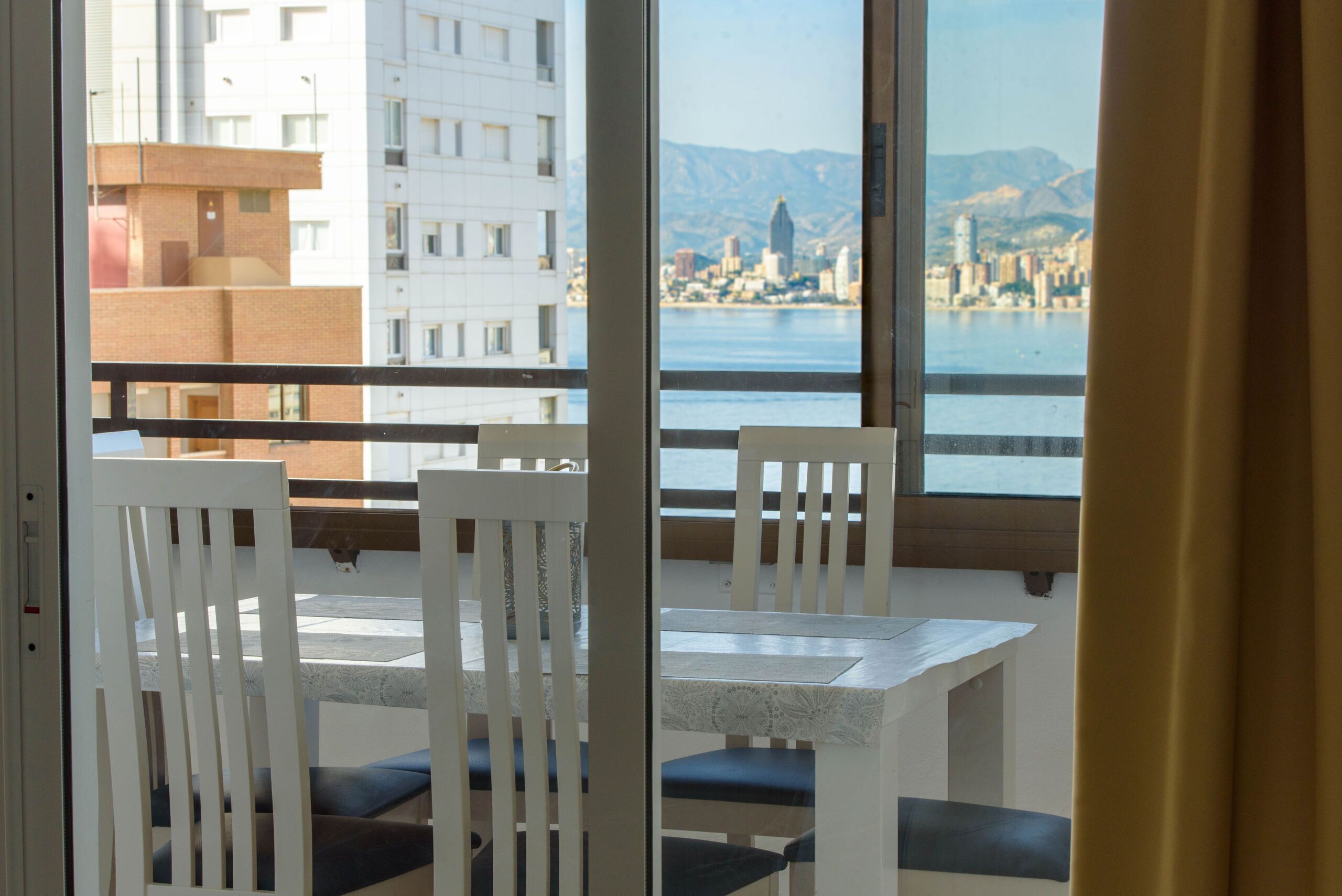 A balcony with white chairs and a table, overlooking a cityscape and water.