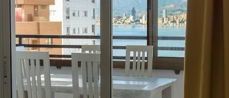 A balcony with white chairs and a table, overlooking a cityscape and water.