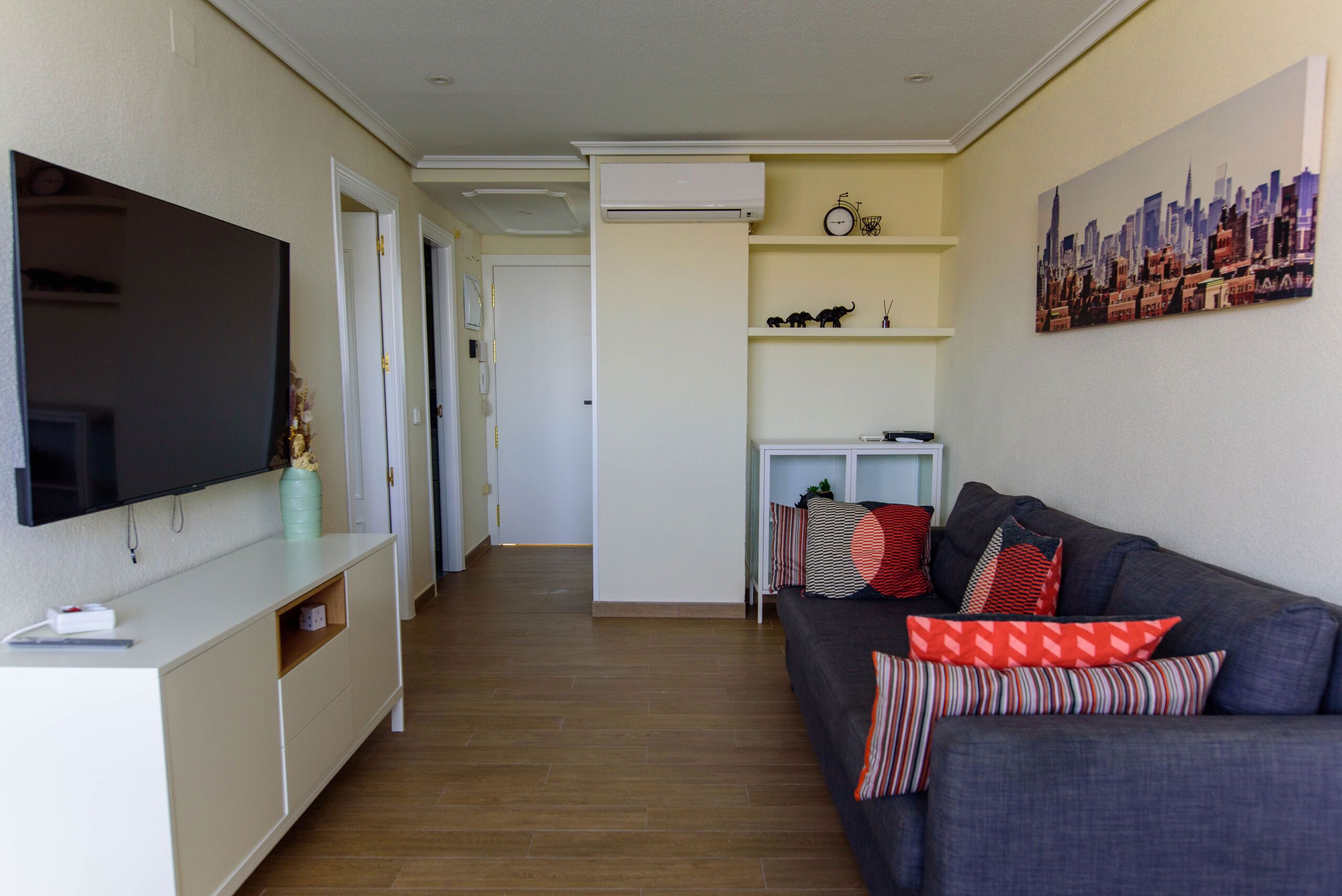 Una sala de estar con un sofá gris, un televisor de pantalla plana, un mueble blanco para equipo audiovisual y un cuadro de una vista urbana en la pared.