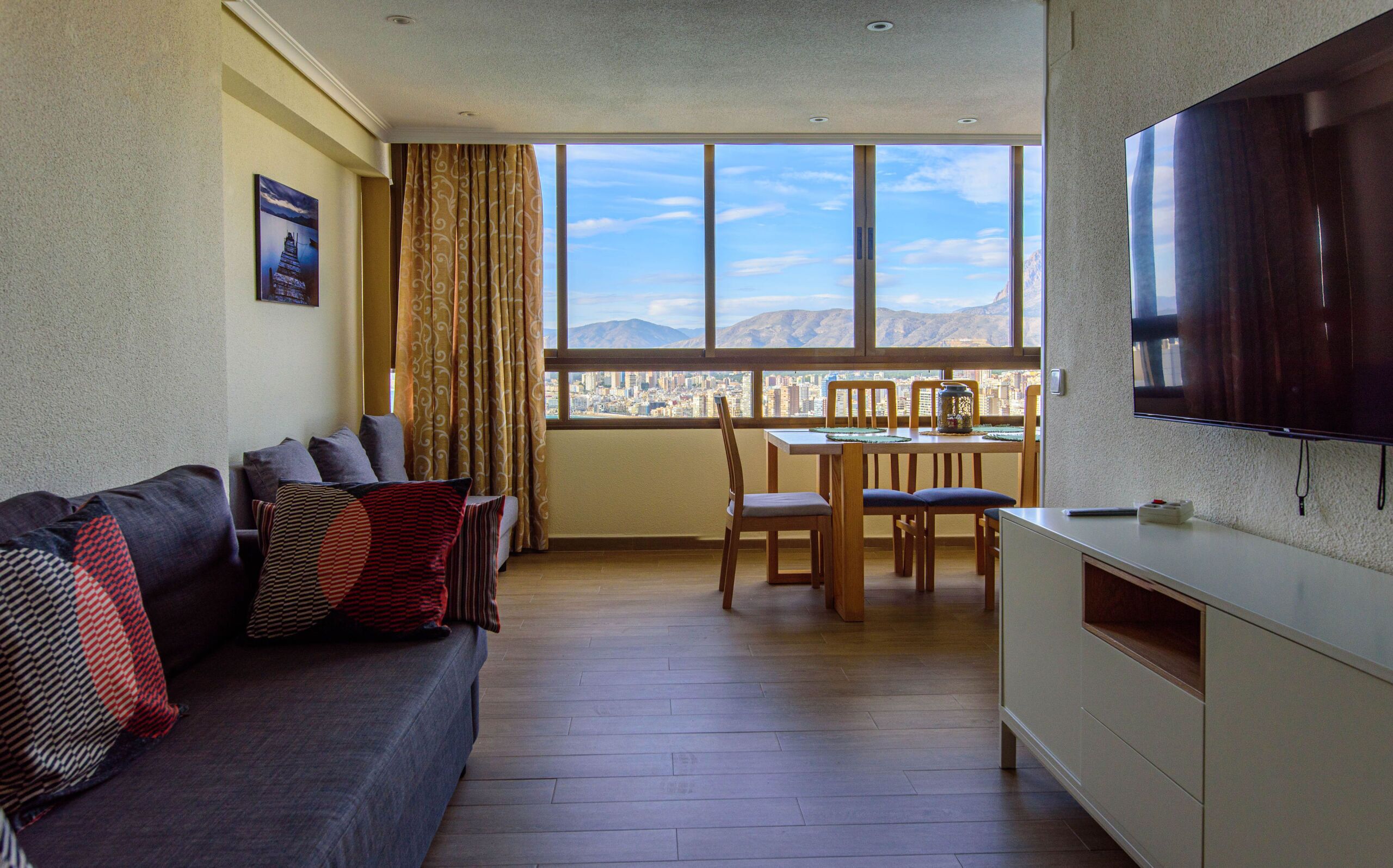 Una sala de estar con sofá, una mesa de comedor con sillas, un televisor montado en la pared y un ventanal con vistas a la ciudad.