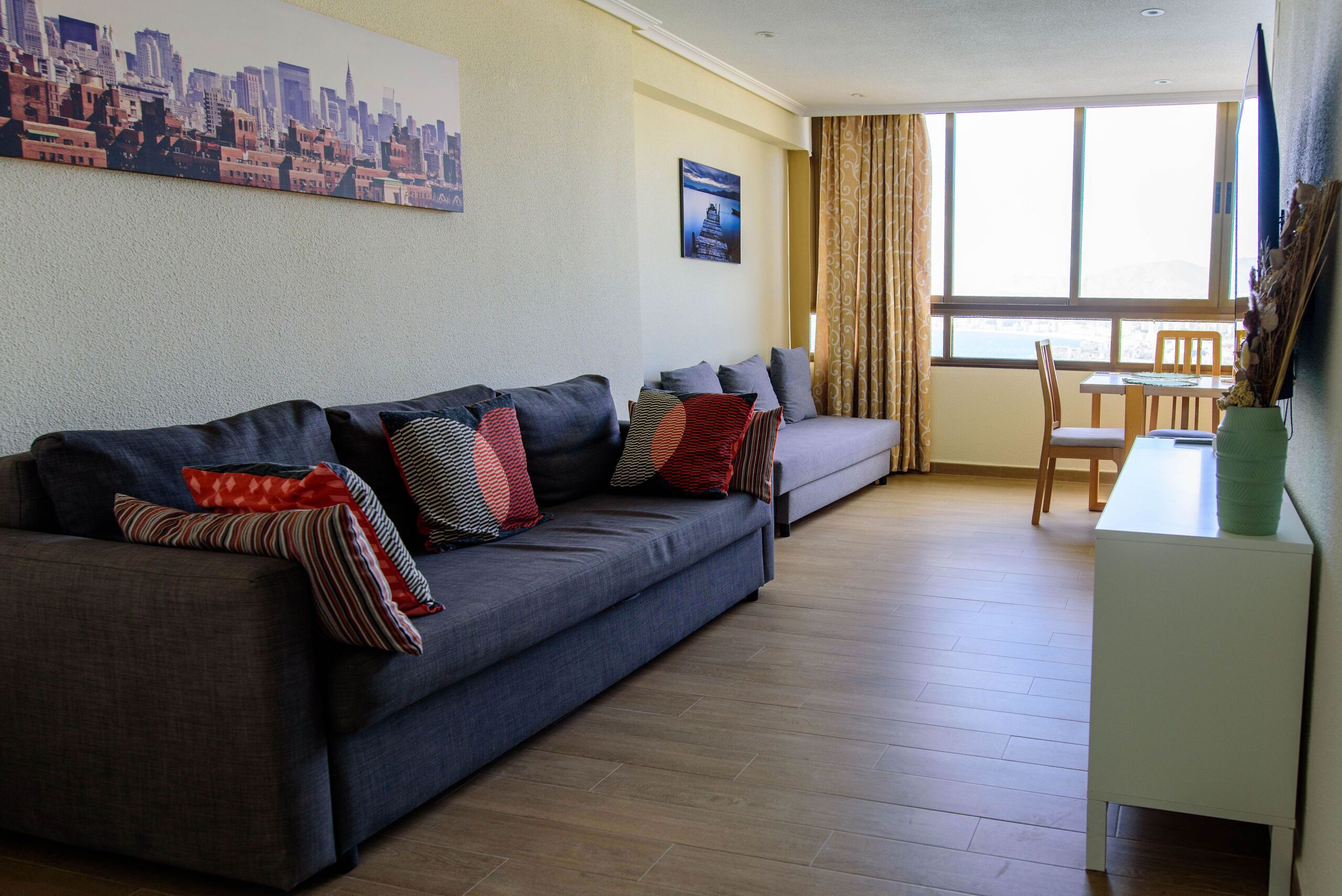 Una sala de estar con sofá, una mesa de comedor y una silla. Hay una ventana con cortinas, un cuadro en la pared y una mesita con un florero.