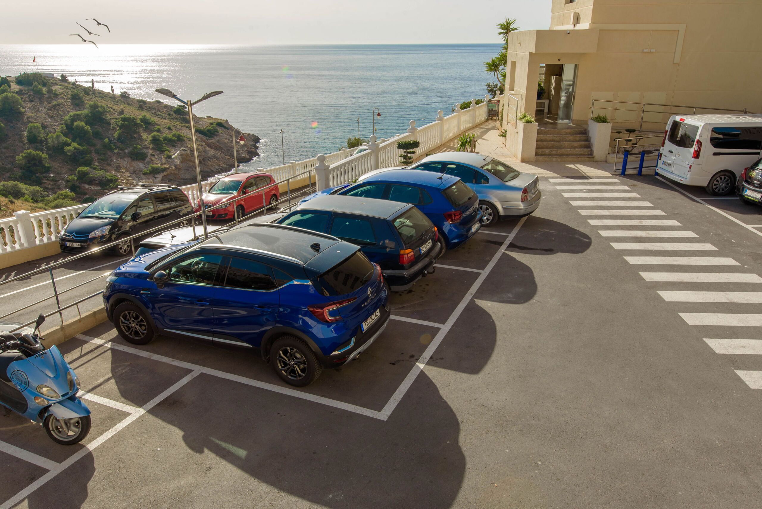 A parking area with multiple cars, a scooter, and a van.