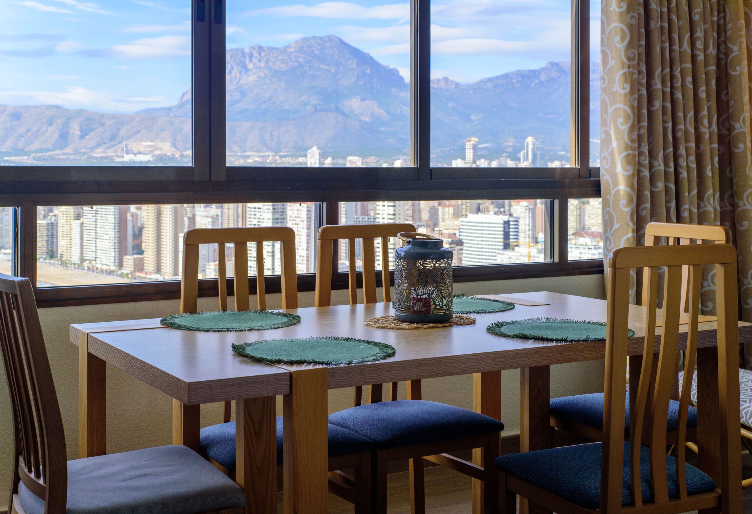 Un coin repas avec une table dressée pour quatre personnes, des chaises dotées de coussins bleus et une vue sur le paysage urbain et les montagnes, visible à travers de grandes fenêtres.