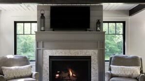 A living room with a fireplace, two armchairs, a flat-screen TV, and a large window with a view of trees.
