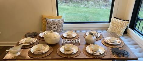 A dining table set with white dinnerware and a pitcher, placed on a wooden bench in front of a window with a view of a grassy yard.