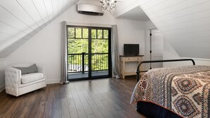 A bedroom with a sloped ceiling, a large window with a view of greenery, a bed with a patterned bedspread, a white armchair, and a desk with a television.