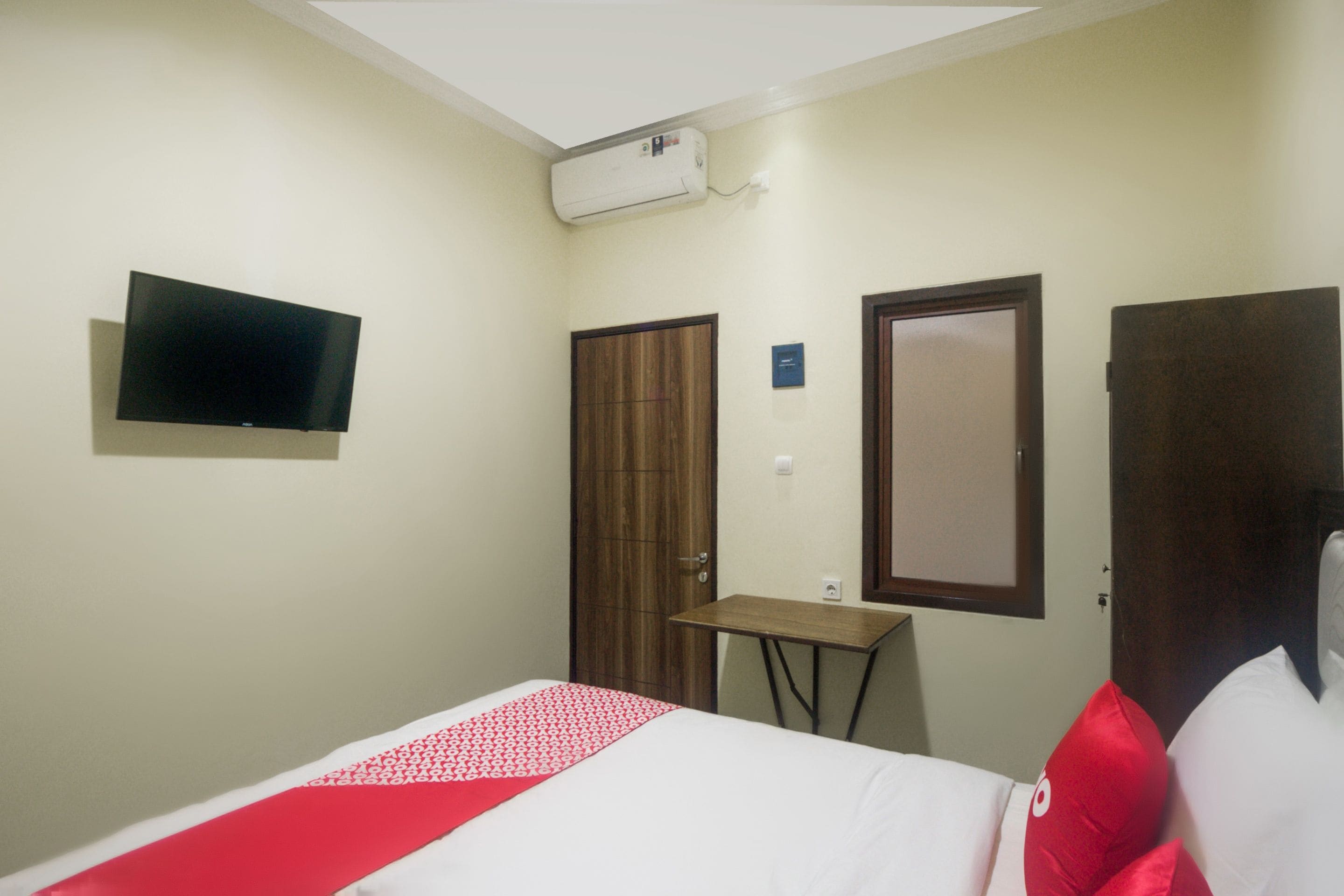 A hotel room with a bed, a TV mounted on the wall, a wooden wardrobe, and a small table.