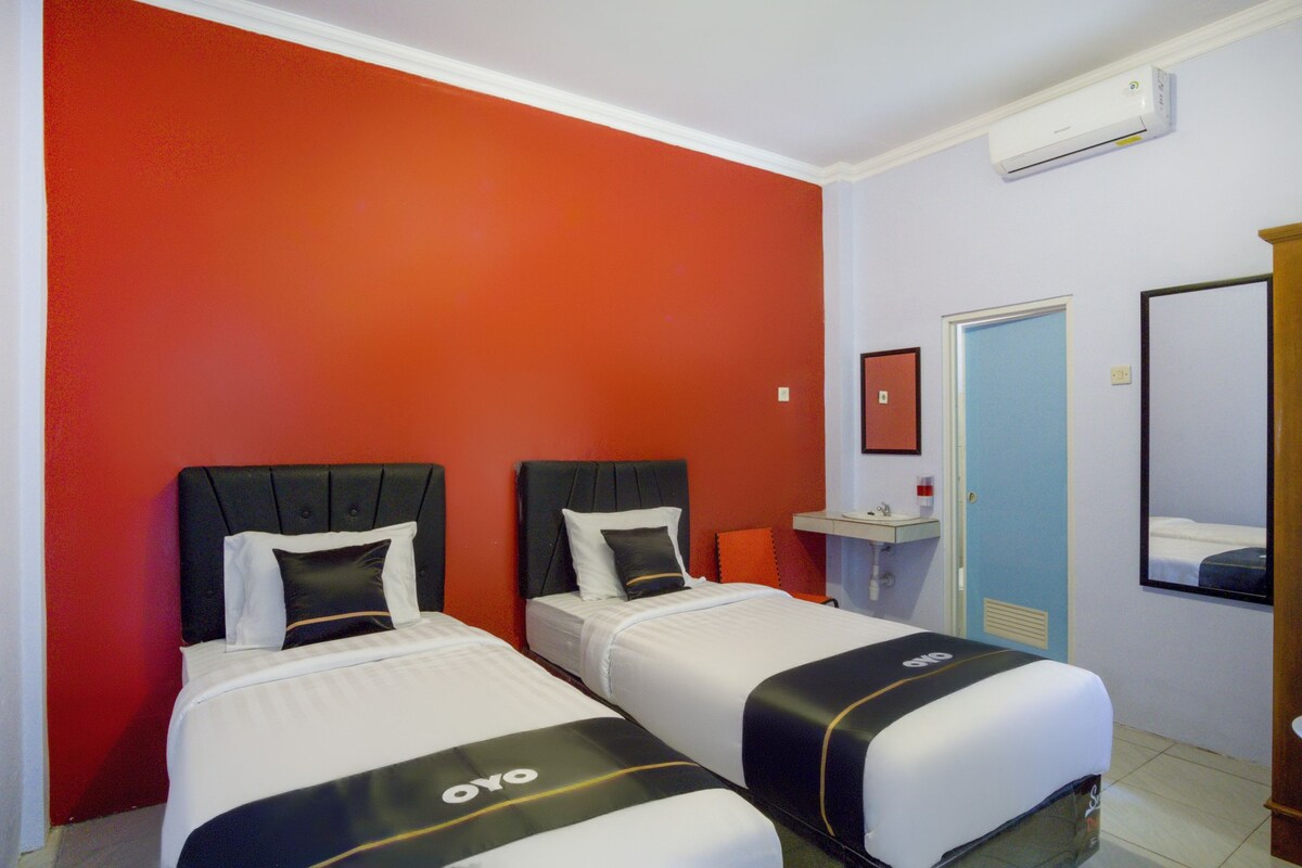 Hotelkamer met twee bedden, een rode accentmuur, een klein nachtkastje met een lamp, een spiegel en een airconditioning.
