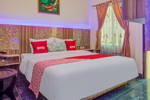 A hotel room with a large bed, two pillows with 'OYO' branding, a red and white patterned throw, a nightstand, a small round table, and a window with curtains.