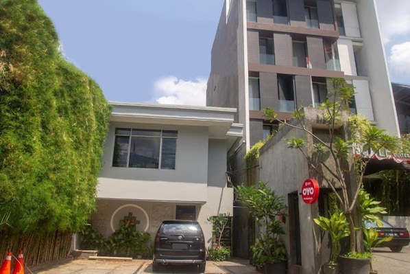 A modern building with a car parked in front, surrounded by greenery and a sign indicating a hotel. - OYO 3931 Wtc Kuningan (Jakarta)